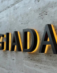 Custom black metal backlit channel letters "GIADA" mounted on a concrete wall with warm halo lighting.
