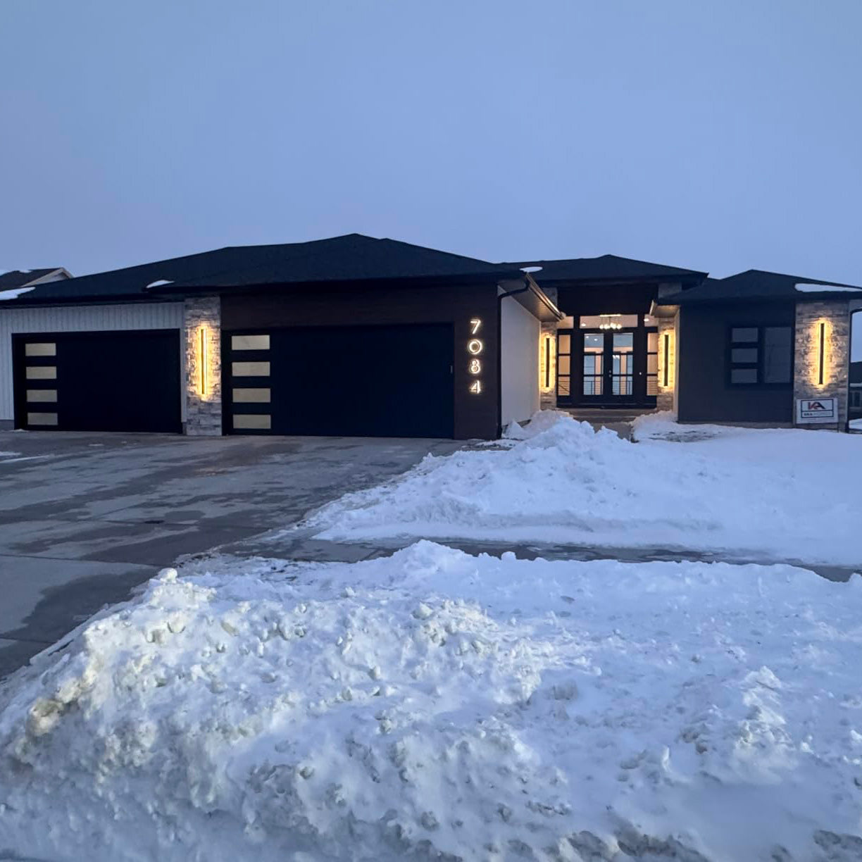 Modern luxury home at night featuring a dark exterior, three lit garage bays, and vertical gold-illuminated address numbers next to the entryway.