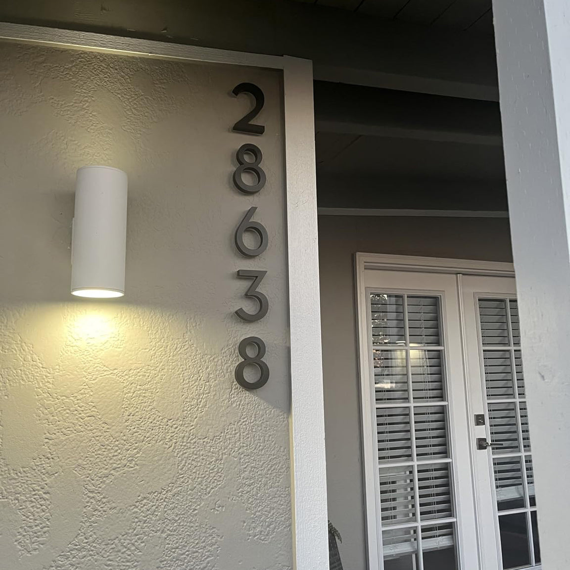 Modern white pillar featuring vertical black illuminated house numbers '28638' and an architectural wall sconce at night.