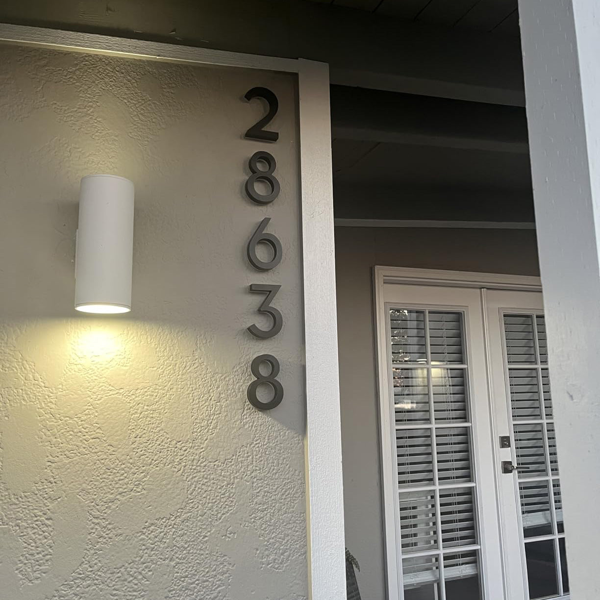 Modern white pillar featuring vertical black illuminated house numbers '28638' and an architectural wall sconce at night.