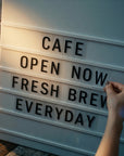 Lightbox with 'CAFE OPEN NOW FRESH BREW EVERYDAY' text on a wooden surface.