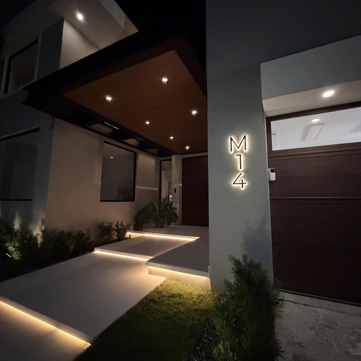 Modern vertical address sign featuring white illuminated 'M14' characters mounted on a gray architectural pillar under a contemporary home entryway at night.