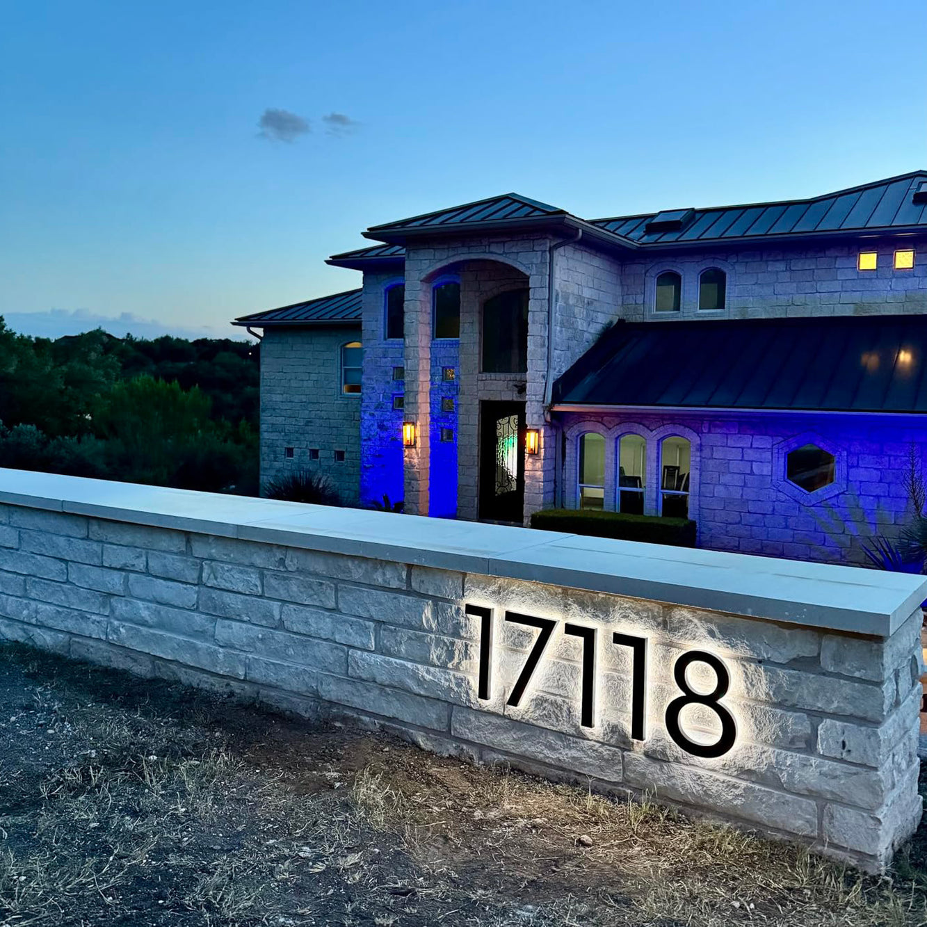 Modern residential address sign with large white illuminated numbers mounted on a gray stone wall at dusk.
