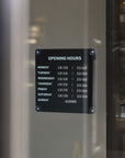 A black metal 'OPENING HOURS' sign with white text, mounted on a glass door or window. The sign lists daily hours from Monday to Sunday as 10:30, with Sunday marked as closed.