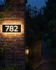 House number '782' on a brick wall at night with a gate in the background.