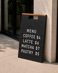 Black A-frame menu board with white letters showing a coffee menu with prices at a storefront entrance