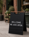 Black A-frame menu board with white letters reading Welcome We Are Open outside a shopfront