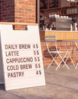 White A-frame menu board with a wood handle showing a coffee menu outside a brick cafe