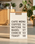 White A-frame menu board with a wood handle showing a cafe menu with prices inside a coffee shop