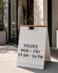 White A-frame menu board with a wood handle showing store hours outside a boutique window