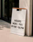 White A-frame menu board with a wood handle showing store hours, leaning on a stone column