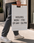 Person carrying a white A-frame hours board one-handed by its wood handle rail along a sidewalk