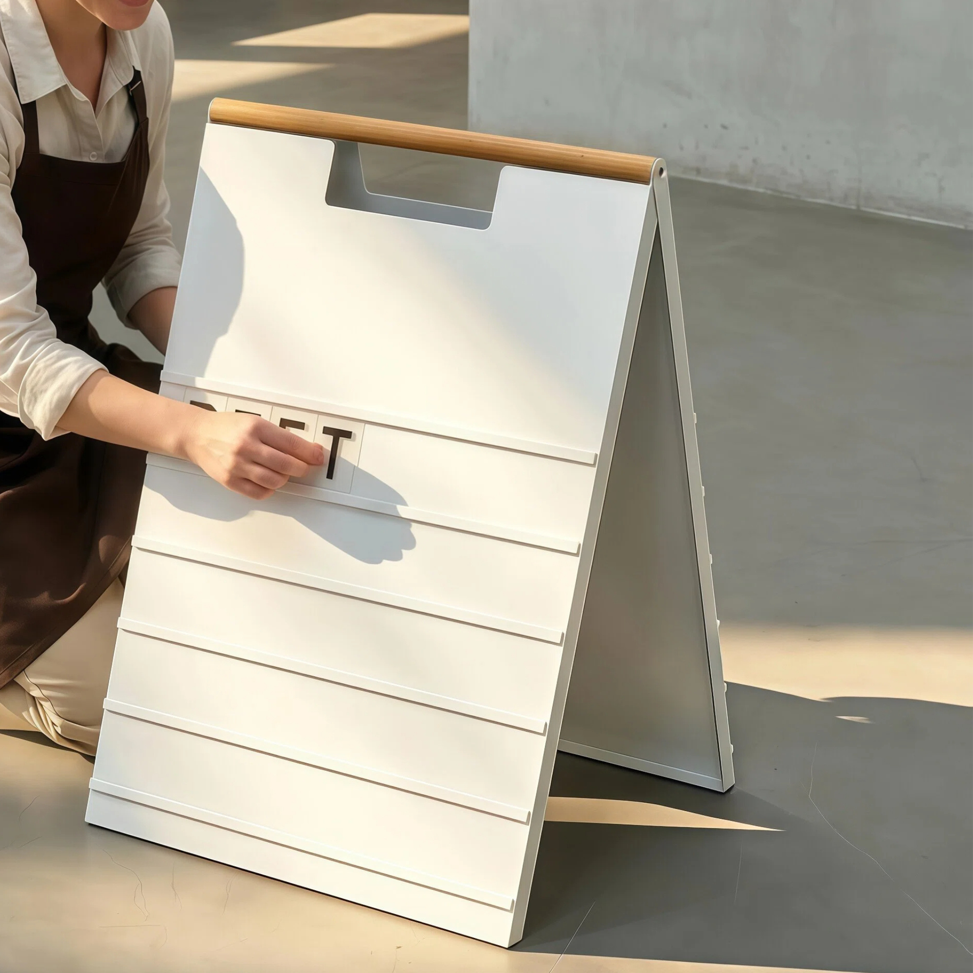 Barista sliding a black letter tile into the rail of a white A-frame menu board with a wood handle