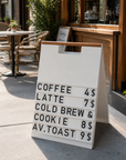 White A-frame menu board with a wood handle showing a coffee price menu on a cafe patio