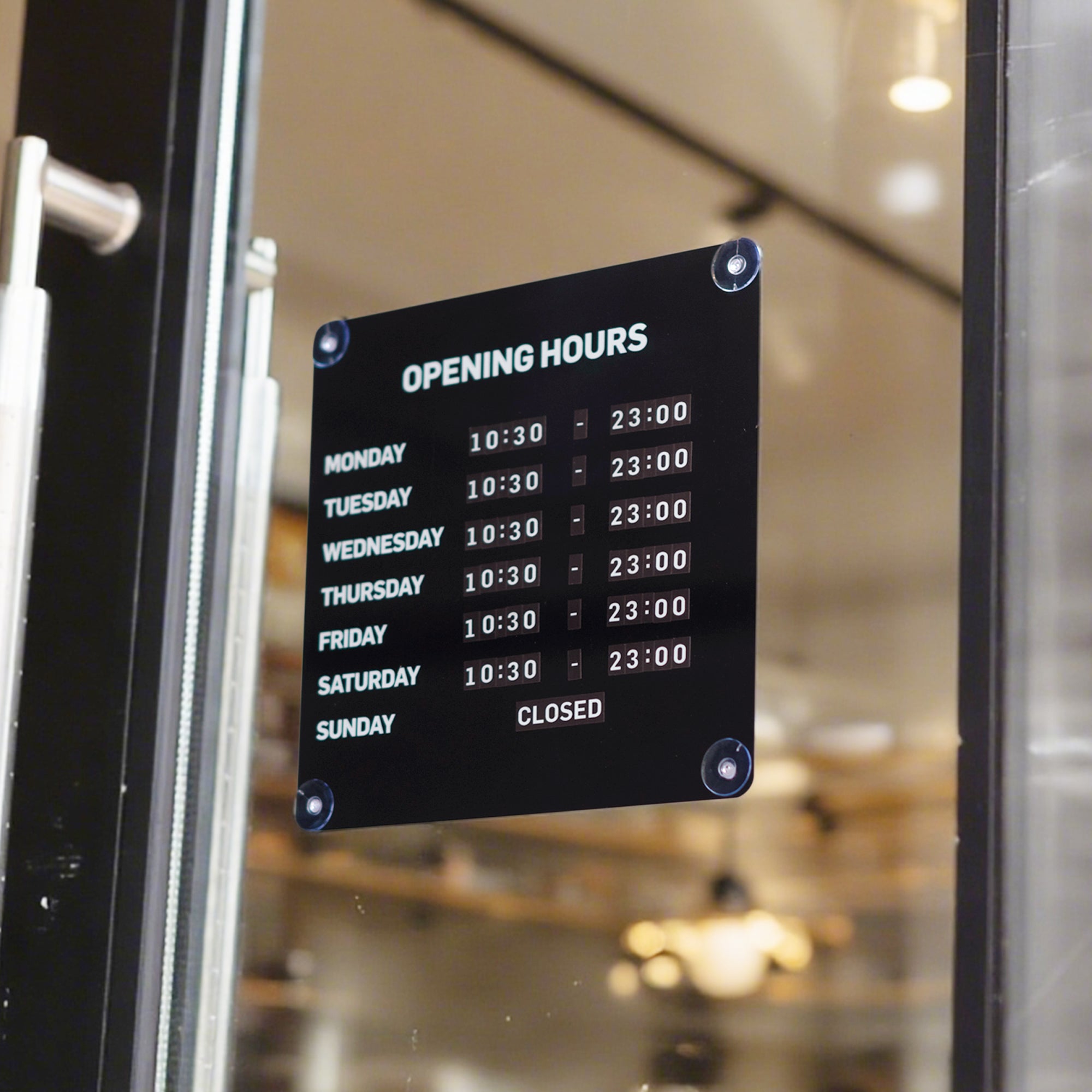 A black rectangular glass door sign displaying opening hours from Monday to Sunday in white text, mounted on a glass door with a blurred indoor shop or cafe background.