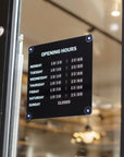 A black rectangular glass door sign displaying opening hours from Monday to Sunday in white text, mounted on a glass door with a blurred indoor shop or cafe background.